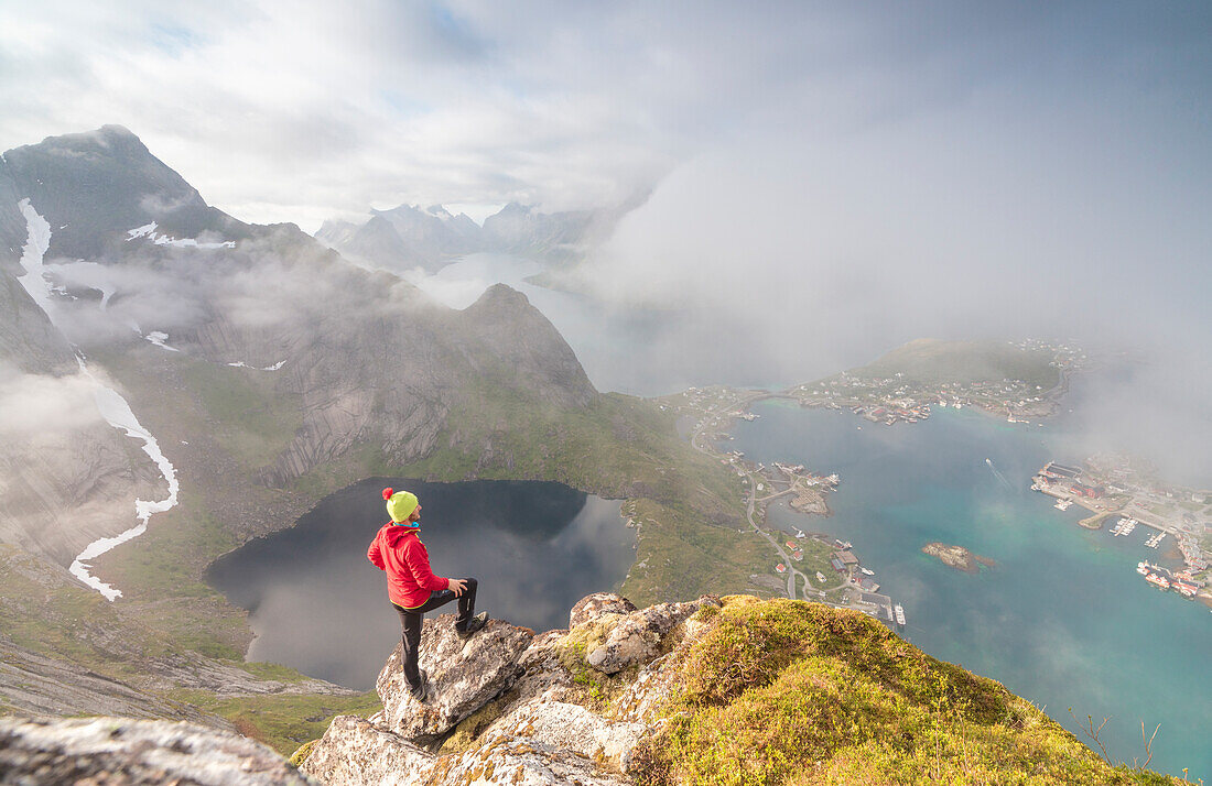 Hiker on summit admires the blue lake and sea framing the village, Reinebringen, Moskenesoya, Lofoten Islands, Norway, Scandinavia, Europe
