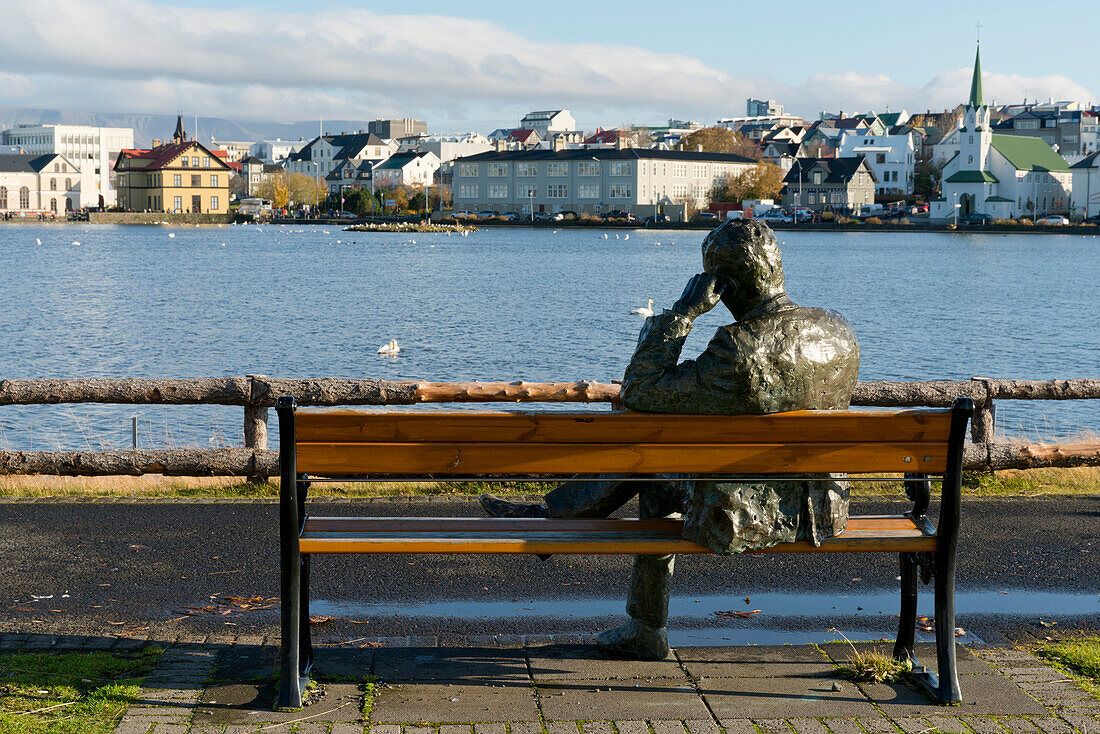 Sculpture of a man sitting on a park bench in front of Tjornin Lake and the Historic Centre of Rykjavik, Iceland, Polar Regions