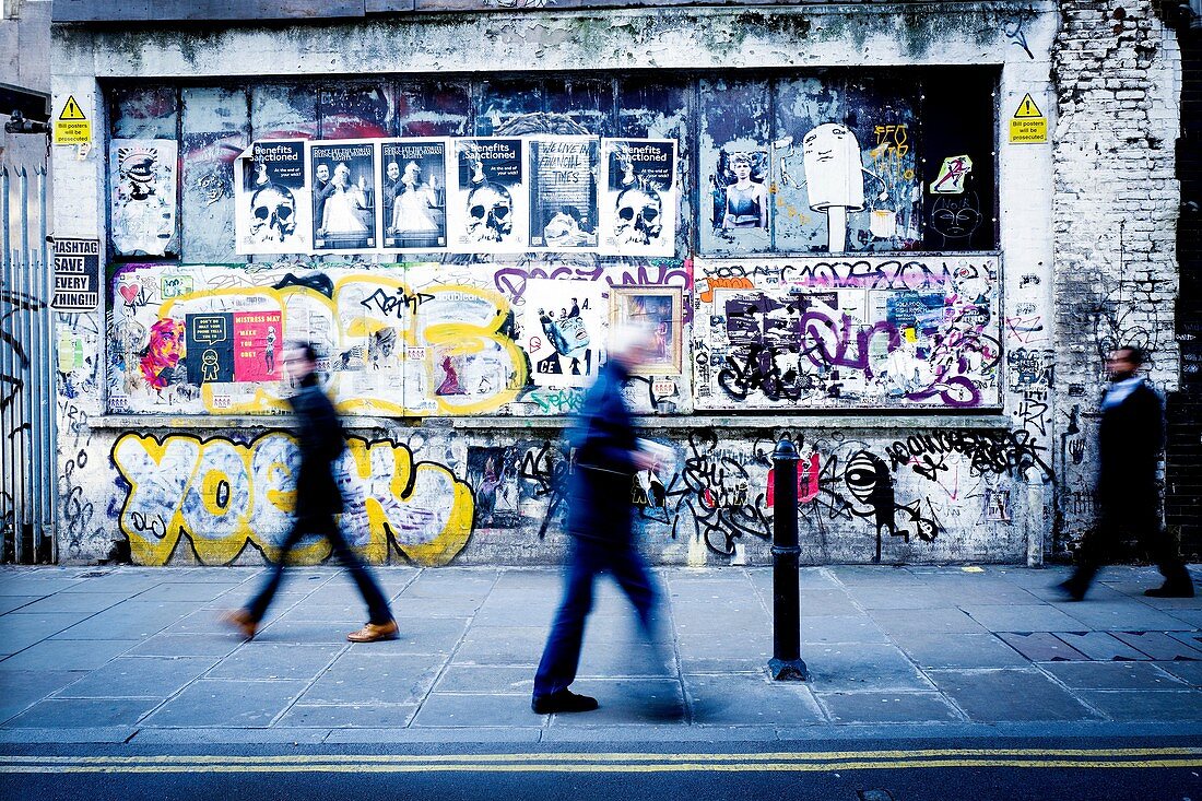 Tres personas irreconocibles andando por la acera de una calle y en la pared de fondo pintada con graffitis. Brick Ln, East End, London, UK, Europa.