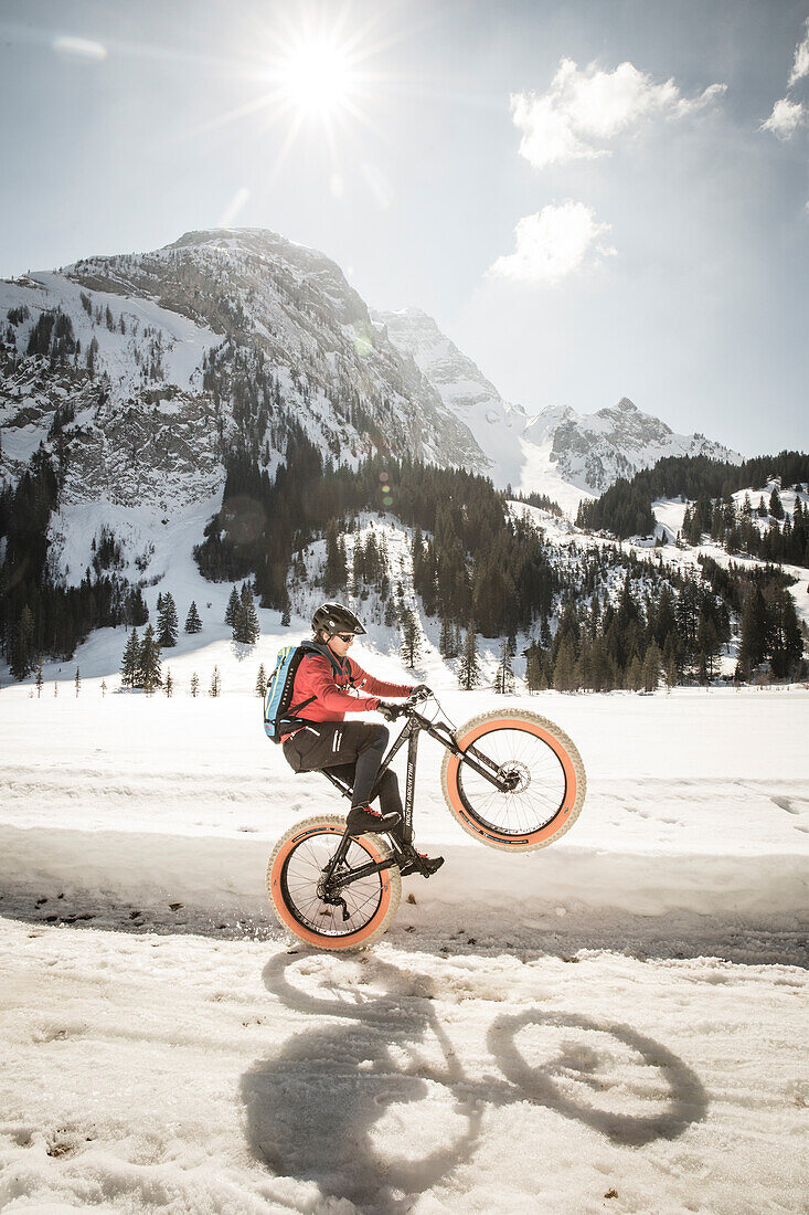 Ein junger Mann zeigt ein Wheelie auf einem Fatbike, Snowbike, Mountainbike vor dem zugefrorenen Lauenensee (Louwenesee) in der Nähe von Gstaad, Berner Oberland, Schweiz