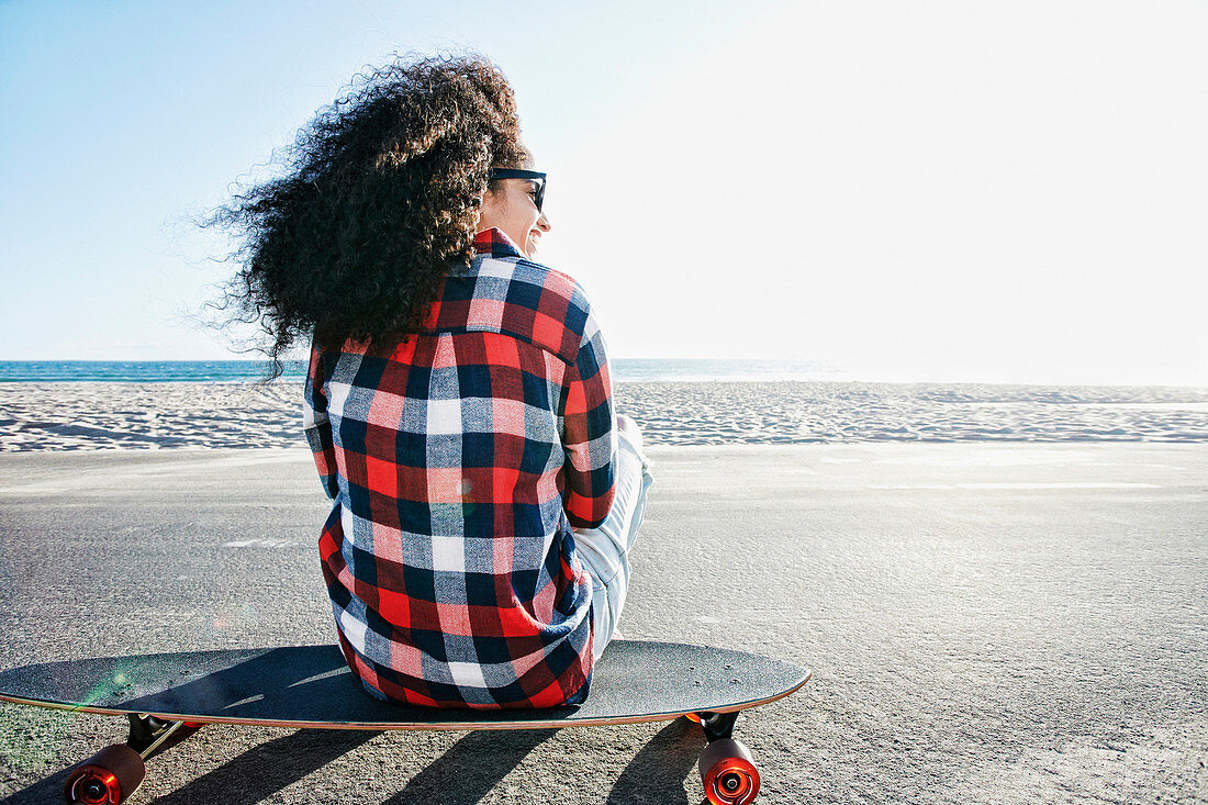 Hispanic woman sitting on skateboard at beach