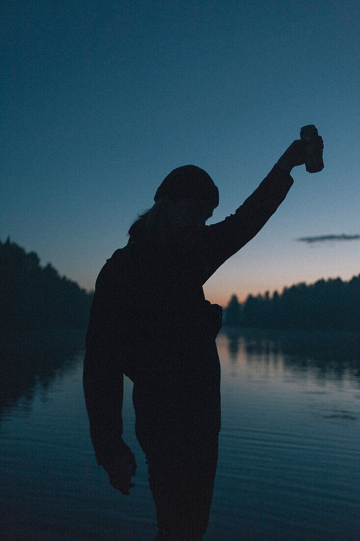 Silhouette of woman holding drink can by lake during sunset