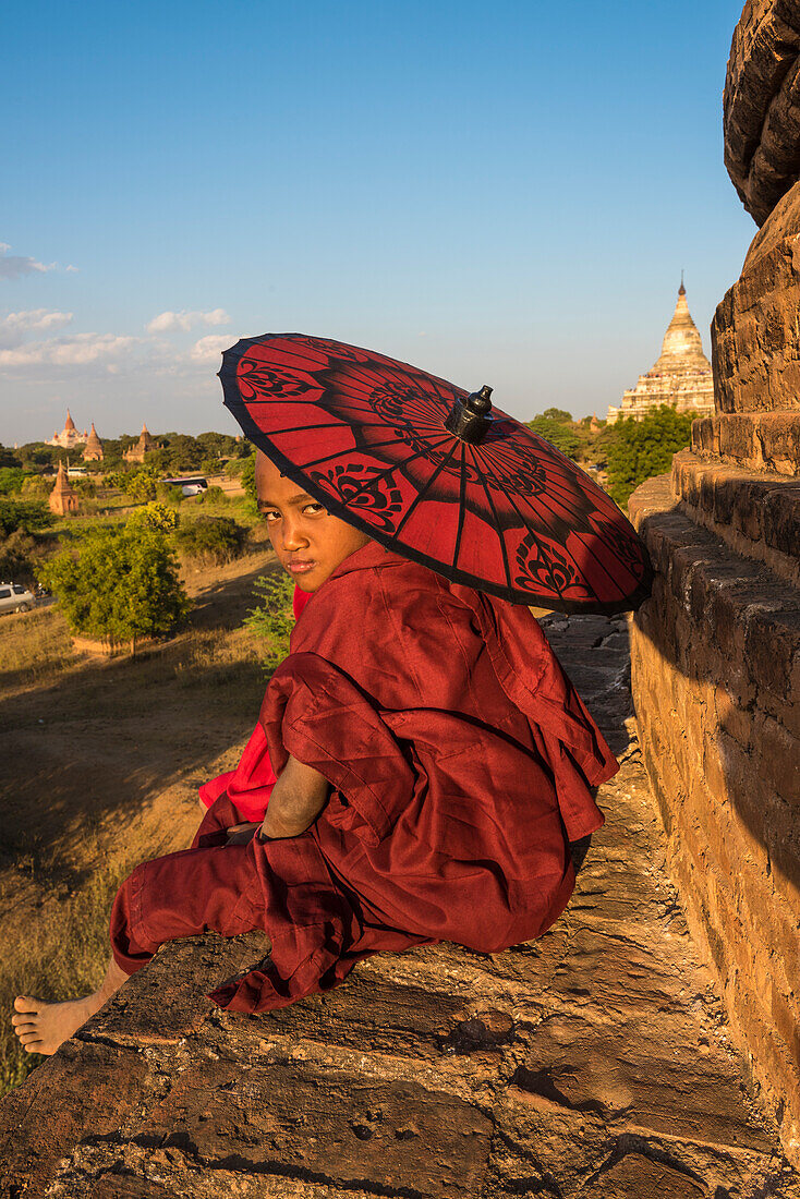 Bagan, Mandalay region, Myanmar Burma , A young monk watching the camera with red umbrella sitting on top of a stupa