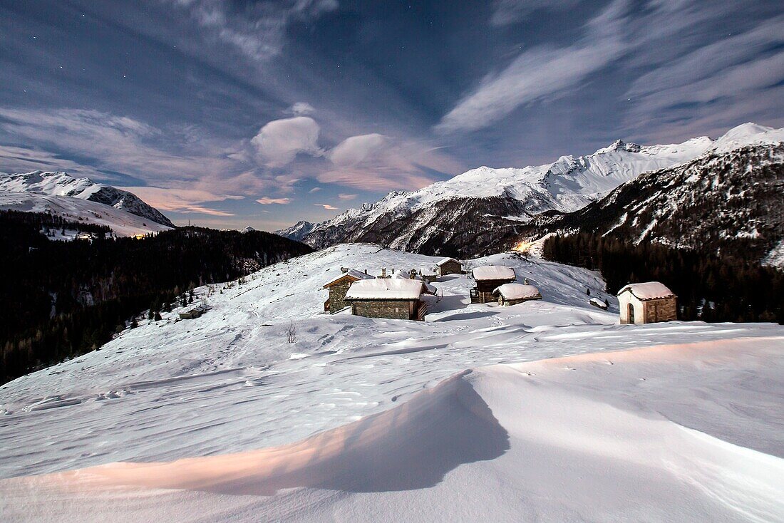 Waves of snow and huts in the small village of Andossi by Madesimo in a full moon night, Valchiavenna, Italy