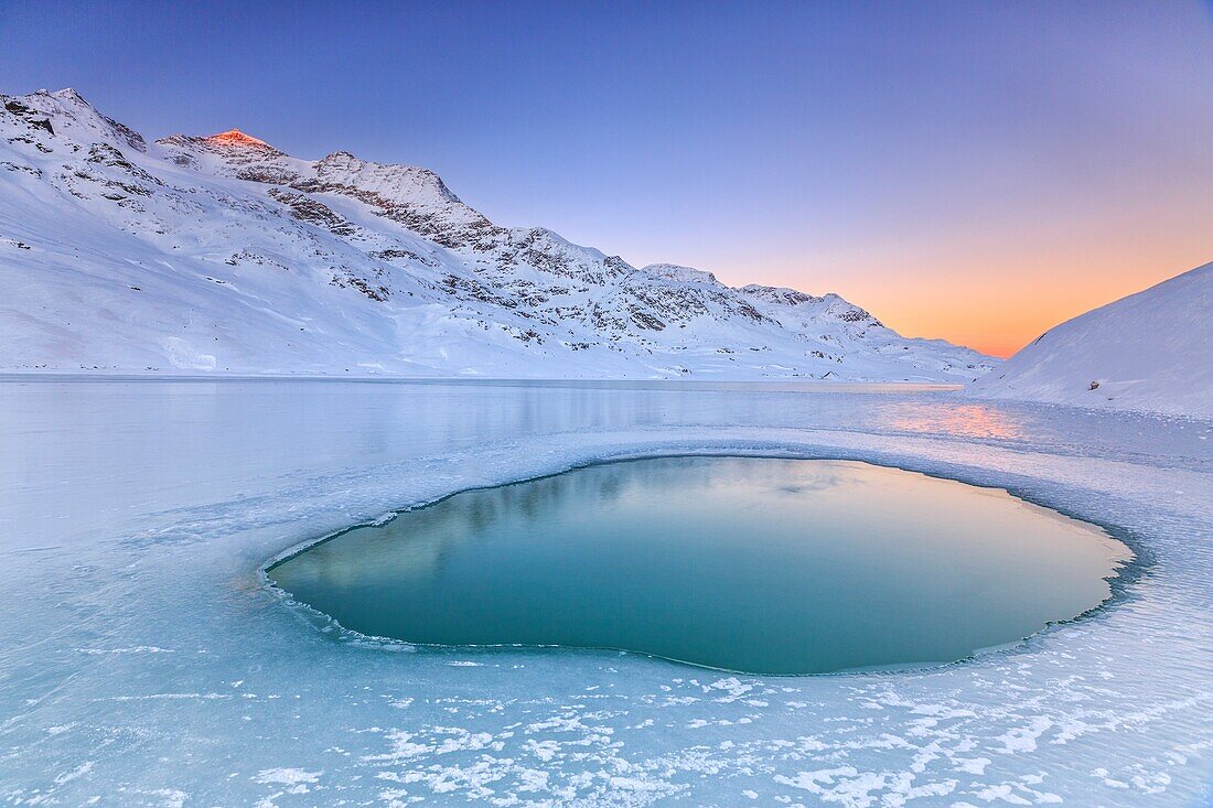 Soft colors of a cold dawn reflected in a pool of turquoise water surrounded by the snow in the middle of White Lake, Bernina Pass, Canton of Graubuenden, Engadine, Switzerland, Europe