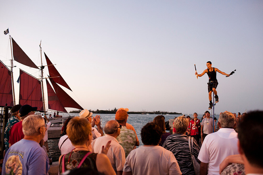 A juggler atop a tall unicycle performs in front of a large crowd of tourists at the famous Key West boardwalk after sunset.