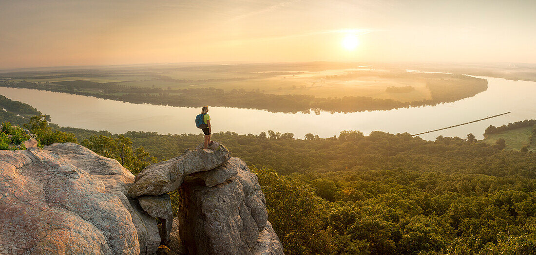 Woman standing on sandstone overhang watching sunrise from summit of Petit Jean Mountain above Arkansas River Valley 
