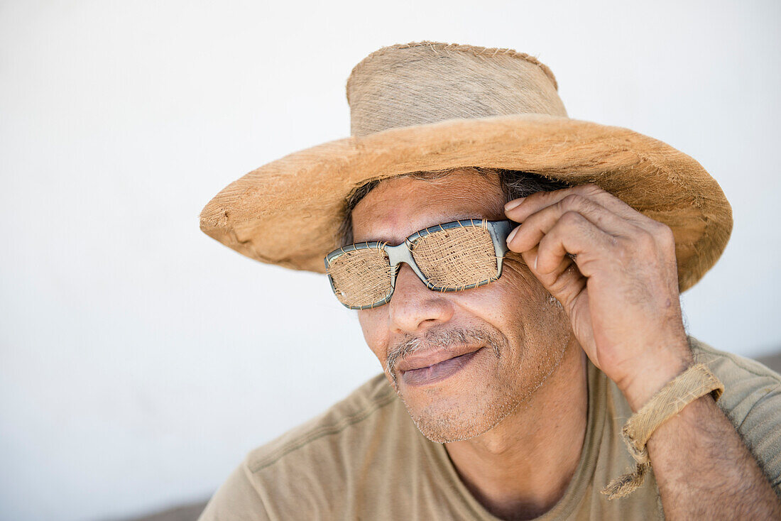 A middle aged mexican man shows off some coconut fibre apparel made by himself, including a hat and sunglasses.