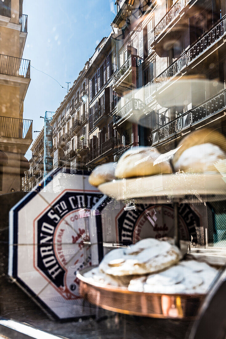 Facades of old houses in the old town are reflected in the shop-window of the baker's Horno Santo Cristo, Palma de Mallorca, Spain