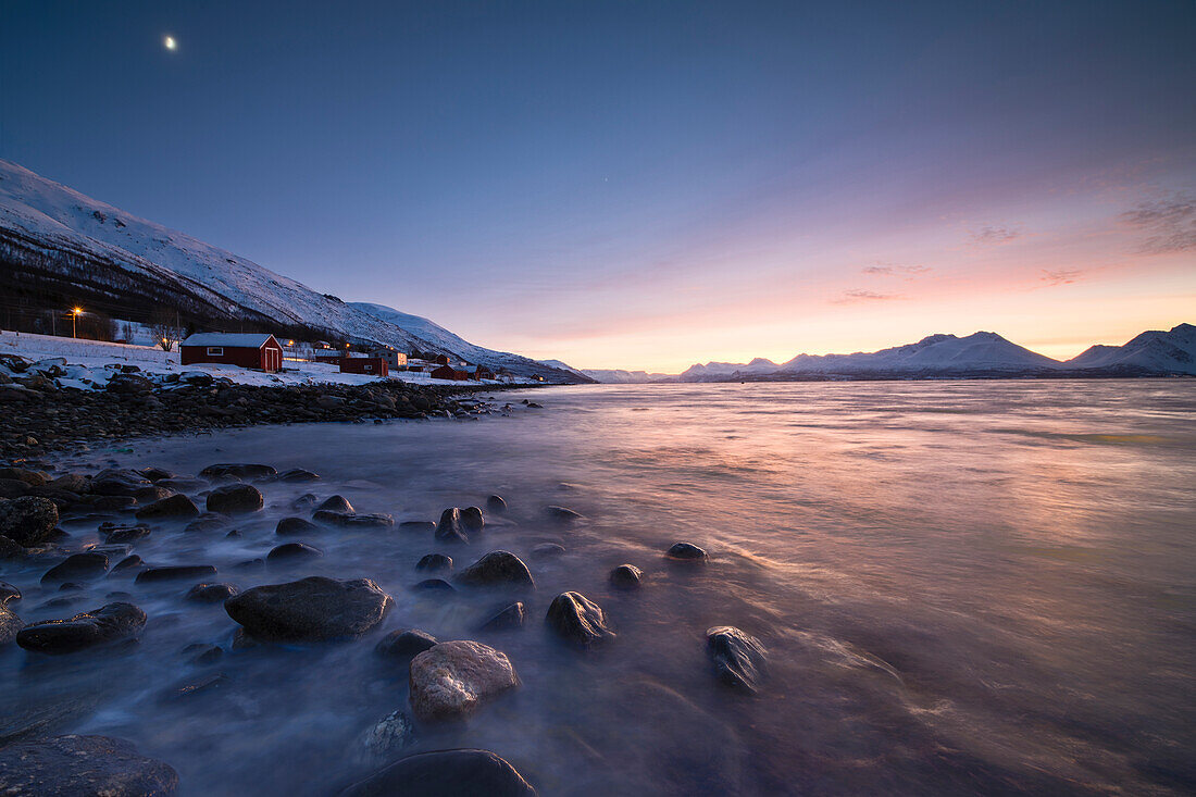 Feuriger Himmel bei Sonnenuntergang auf typischen Rorbu umrahmt von schneebedeckten Gipfeln und gefrorenem Meer, Djupvik, Lyngen Alpen, Troms, Norwegen, Skandinavien, Europa