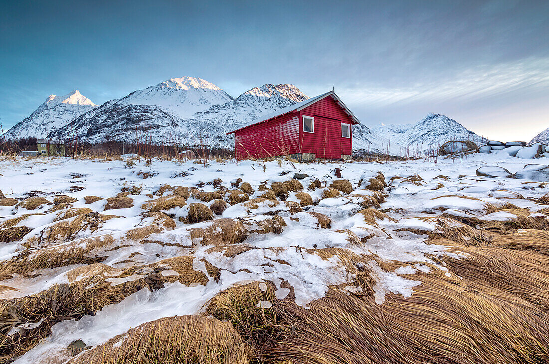 Holzhütte umrahmt von Felsen bedeckt mit Gras und Eis mit schneebedeckten Gipfeln im Hintergrund, Svensby, Lyngen Alpen, Troms, Norwegen, Skandinavien, Europa