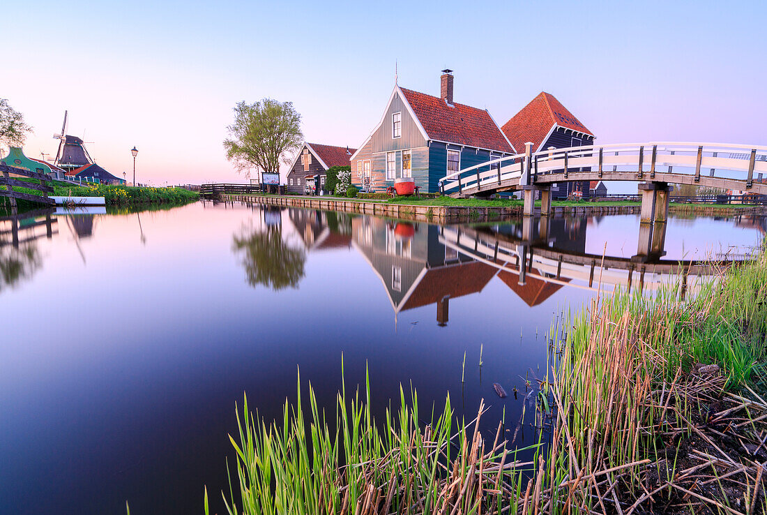 Holzhäuser und Windmühle spiegelt sich im blauen Fluss Zaan bei Sonnenuntergang, Zaanse Schans, Nordholland, Niederlande, Europa