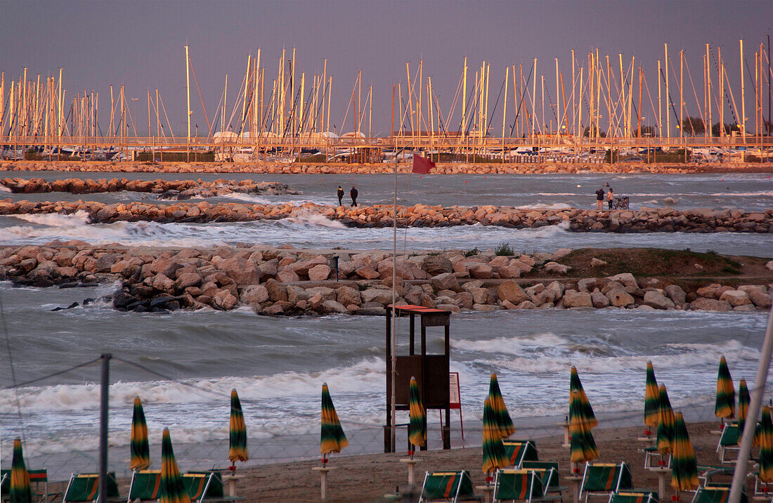 Der Sonnenuntergang am Rimini-Strand beleuchtet die rauen Meere und den kleinen touristischen Hafen mit verankerten Segelbooten auf den Docks von Steinen, Emilia Romagna, Italien