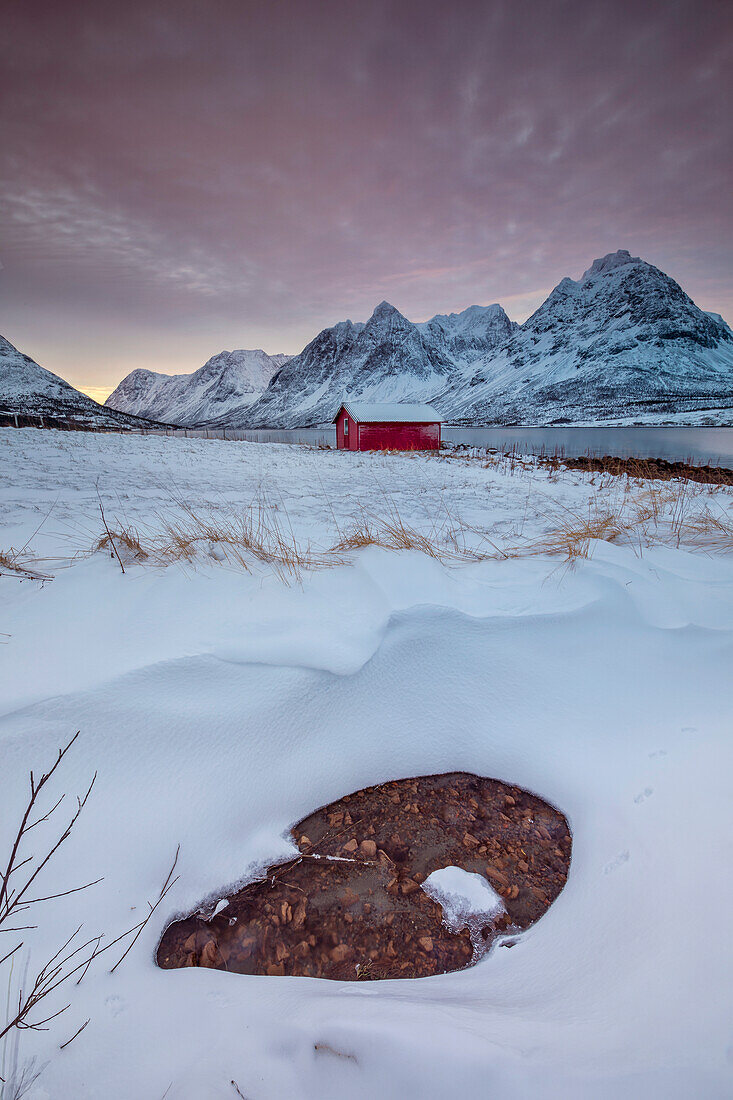 Dark clouds at dawn on the wooden hut surrounded by frozen sea and snowy peaks Svensby Lyngen Alps Tromsø Norway Europe