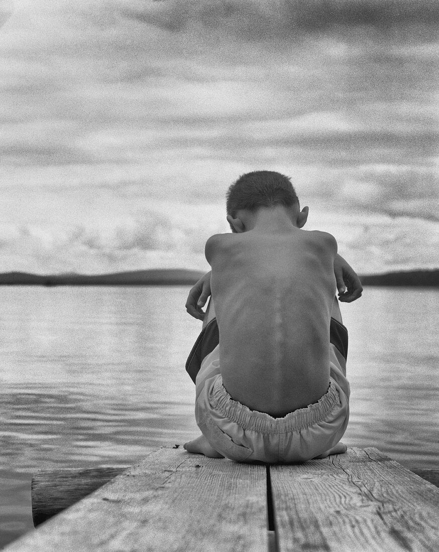 Rear View Portrait of Sad Boy Sitting Hugging His Knees at End of Dock on Lake