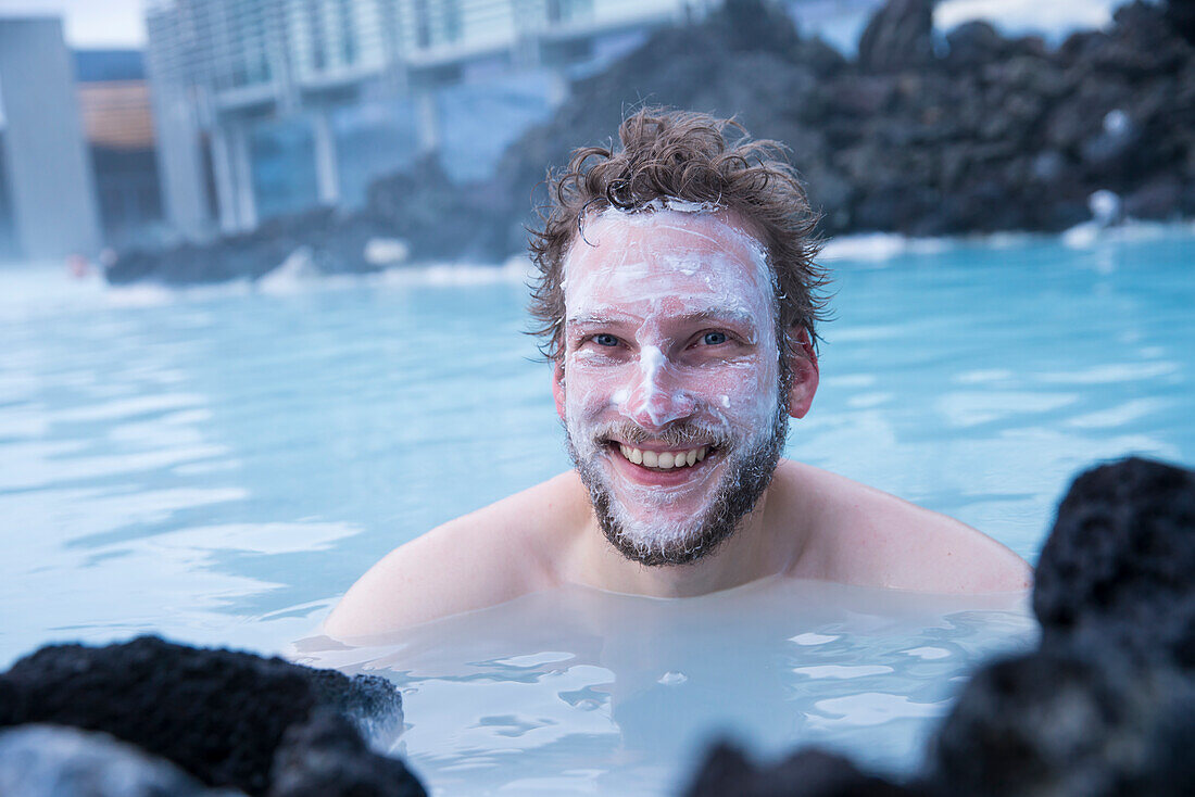 Young man wears silica mud mask and smiles while bathing at The Blue Lagoon, the famous wellness thermal pool with its typical blue-white water in winter time, near Grindavik, Reykjanes, Iceland, Europe