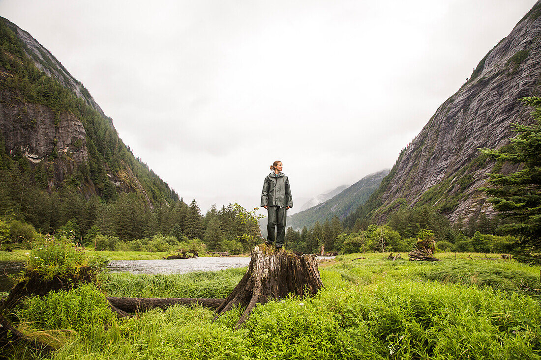 A woman a green rain coat and pants stands on a giant stump in a grassy valley with steep cliff walls.
