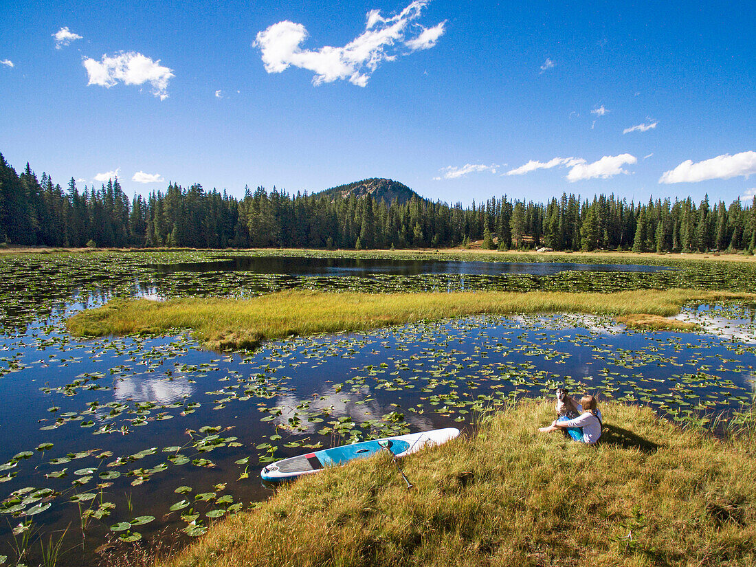 Young female sit with dog along an alpine lake with her paddleboard on a sunny afternoon