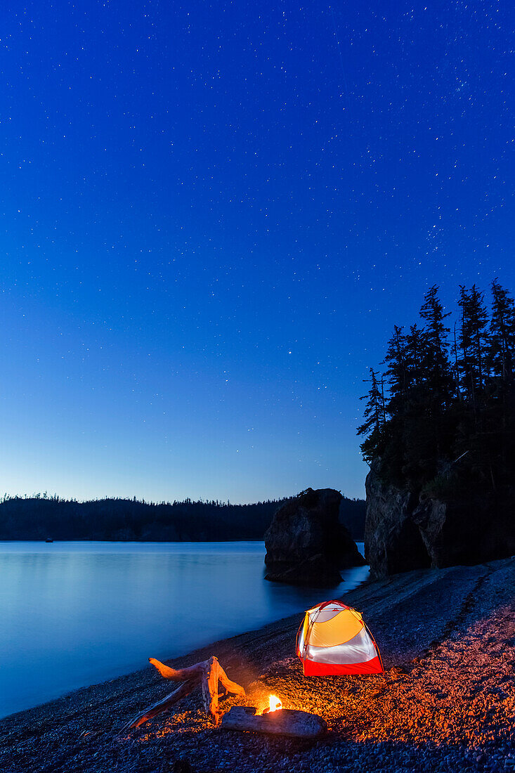 'A campfire built on a beach at dusk next to a glowing tent, tranquil ocean water reflecting the warm light, Hesketh Island; Homer, Alaska, United States of America '