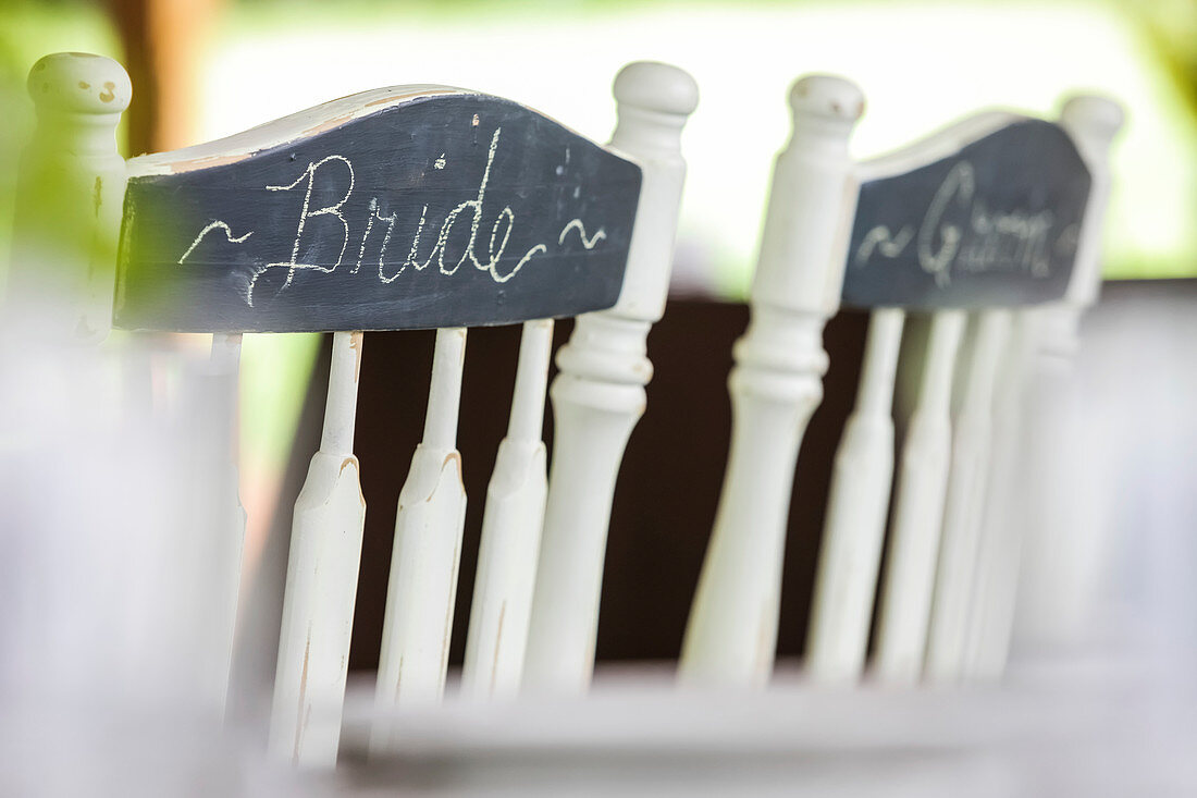Weiße hölzerne Stühle nebeneinander mit Braut und Bräutigam geschrieben in Kreide auf einer Tafel auf dem Rücken