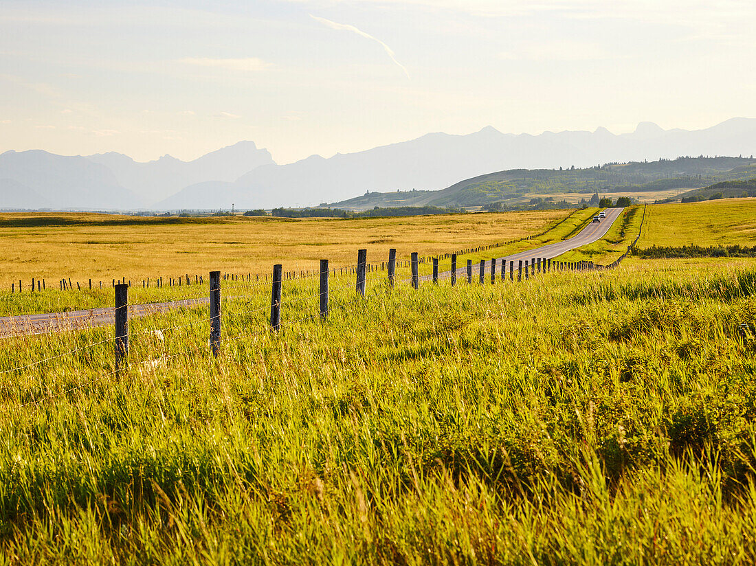 'Eine Landstraße zwischen grünen Feldern und einer Silhouette der Berge in der Ferne; Cochrane, Alberta, Kanada'
