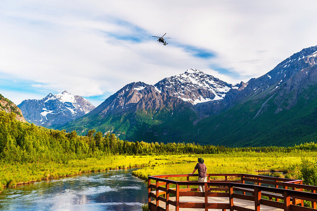 A man is standing on the Eagle River Nature Center boardwalk while a Black Hawk helicopter flies over head in the Chugach State Park in Southcentral Alaska, USA