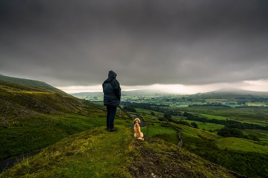 'A man stands with his dog on a grassy hill looking out over the lush, green landscape under a stormy sky; North Yorkshire, England'