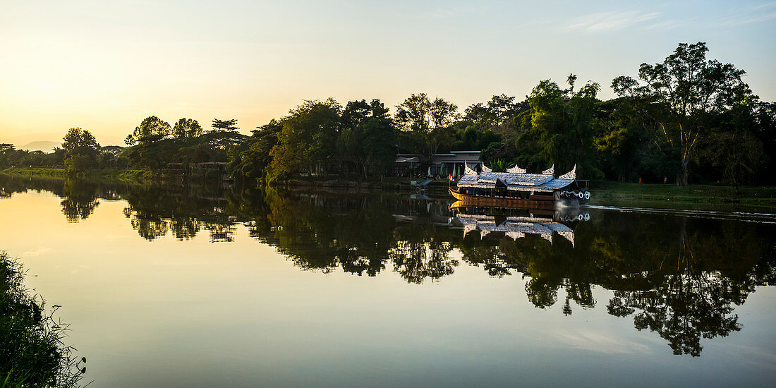 'Reflections in a tranquil river at sunset with a tour boat along the shoreline; Chiang Rai, Thailand'