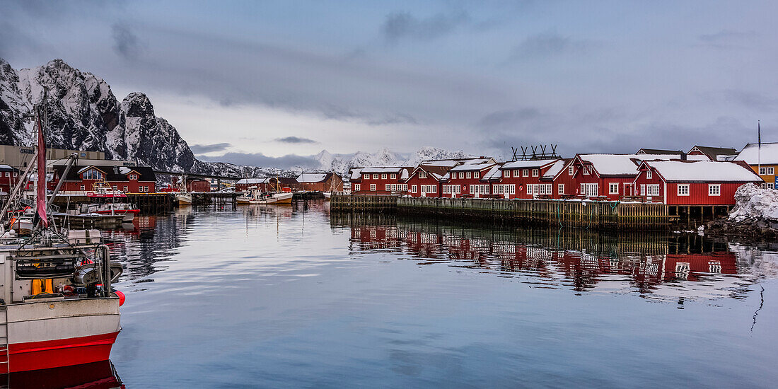 'Red buildings line the water's edge and reflect into the tranquil water with boats moored along the shoreline; Svolvar, Lofoten Islands, Nordland, Norway'