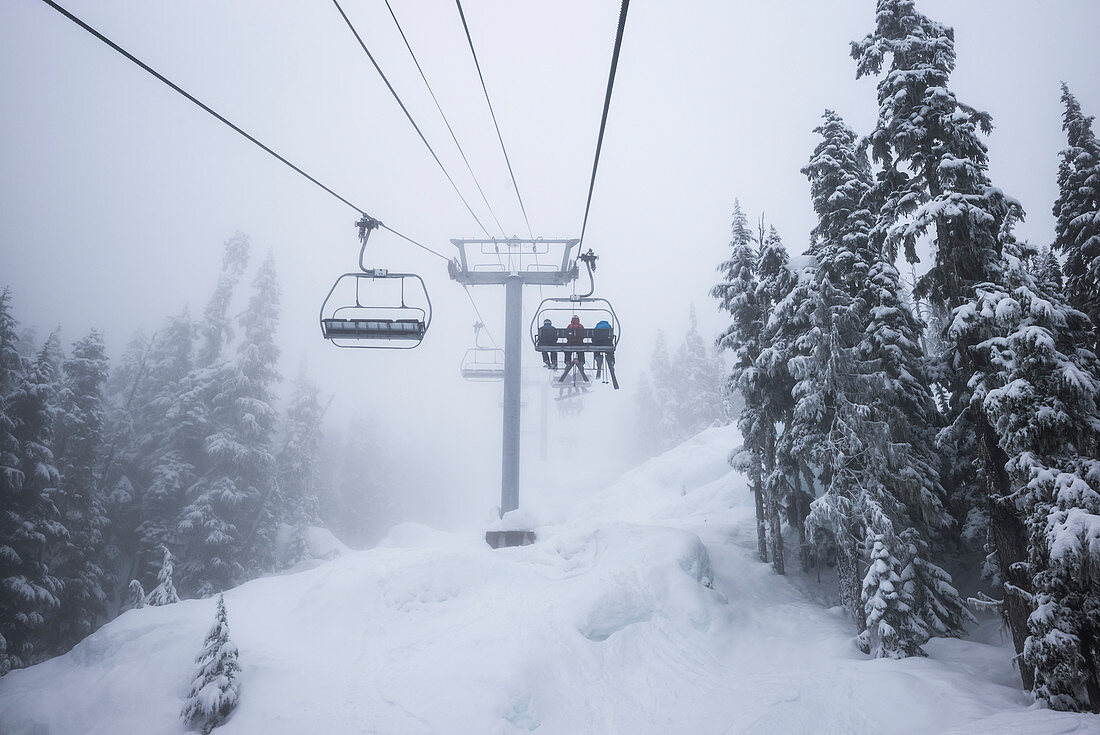 'Downhill skiiers ride the chairlift at a ski resort in the blowing snow and cloud; Whistler, British Columbia, Canada'