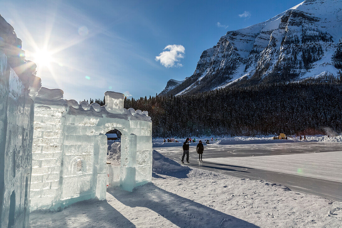 'The sun shines behind an ice castle constructed on frozen Lake Louise in Banff National Park with figure skaters skating on the lake in the background; Lake Louise, Alberta, Canada'