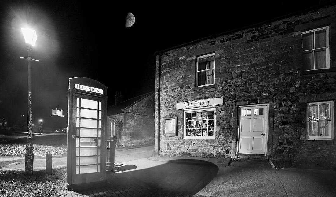 'Eine Telefonzelle, die von einer Straßenlaterne nachts außerhalb eines Geschäftes beleuchtet wird; Bamburgh, Northumberland, England'