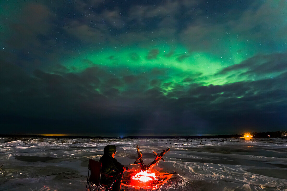 Ein Mann erwärmt sich neben einem Treibholzfeuer, während er die Aurora durch Wolken am gefrorenen Delta River, in der Nähe von Delta Junction, Alaska, beobachtet