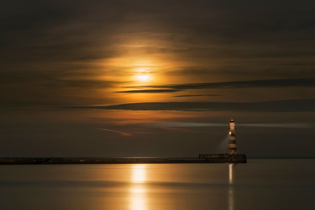 Roker lighthouse casts a light with the golden sunlight shining through cloud and reflecting on tranquil water, Sunderland, Tyne and Wear, England