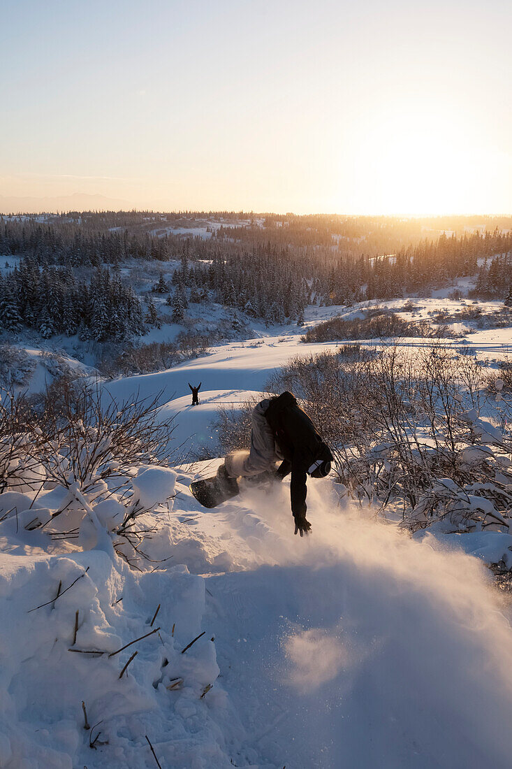 Snowboarder in mid-air with a friend cheering with arms raised on a hill below, Alaska, United States of America