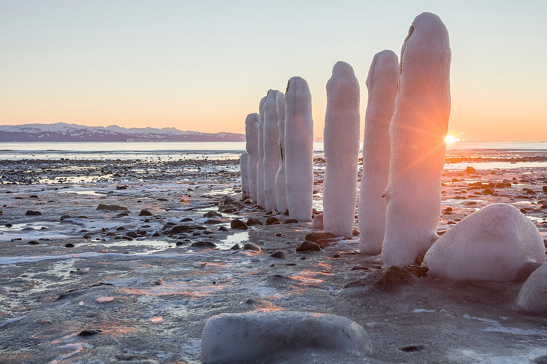 Felsen und Pfähle, die mit Schnee und Eis am Ufer des Wassers bedeckt sind, mit Blick auf die Küste und die Berge in der Ferne, Alaska, Vereinigte Staaten von Amerika