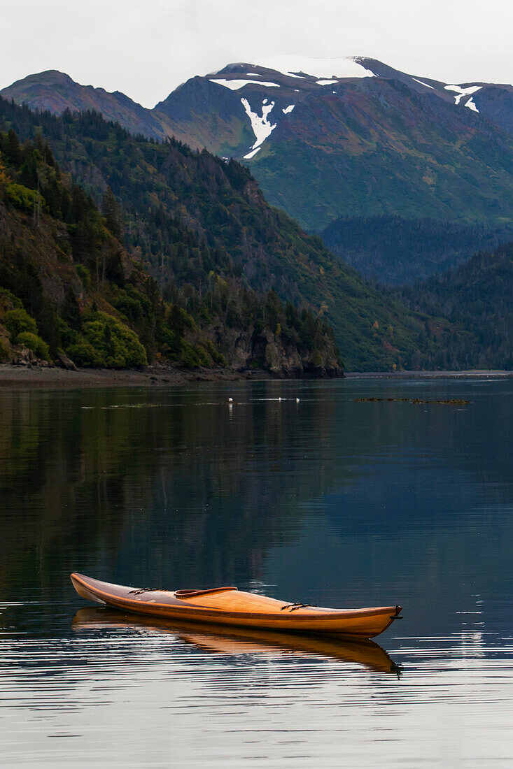 Ein Kajak sitzt auf dem ruhigen Wasser mit Blick auf Wälder und eine Bergkette im Hintergrund, Kachemak Bay State Park, Alaska, Vereinigte Staaten von Amerika