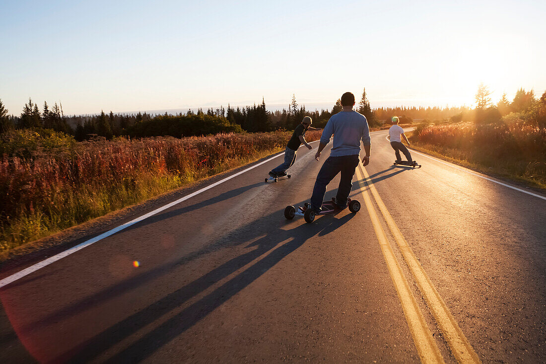 Drei junge Männer skaten eine Straße bei Sonnenuntergang hinunter, Homer, Alaska, Vereinigte Staaten von Amerika