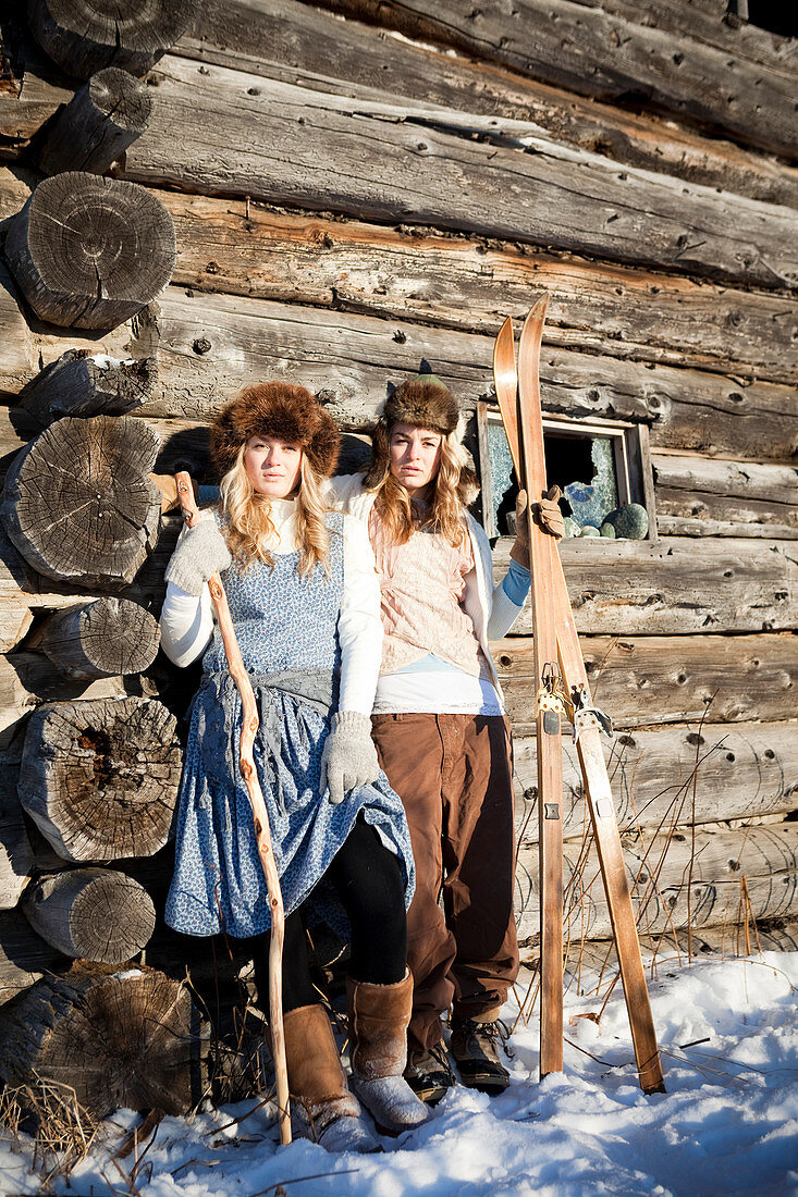 Portrait of two girls standing by a log cabin wearing fur hats holding skis and a walking stick, Homer, Alaska, United States of America