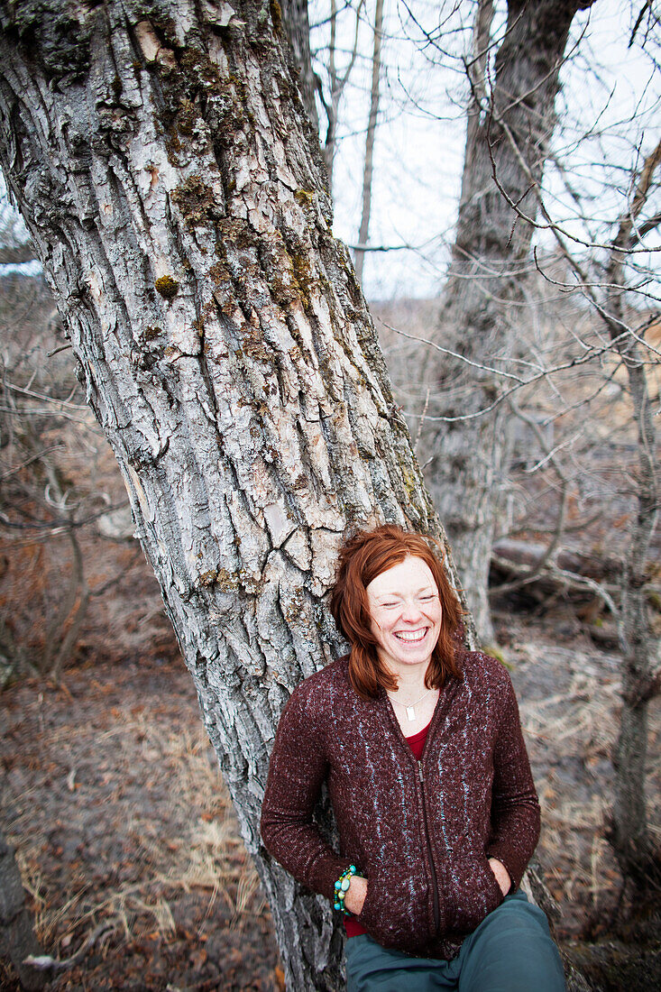Portrait of a woman with red hair leaning on a tree and laughing with eyes closed, Homer, Alaska, United States of America