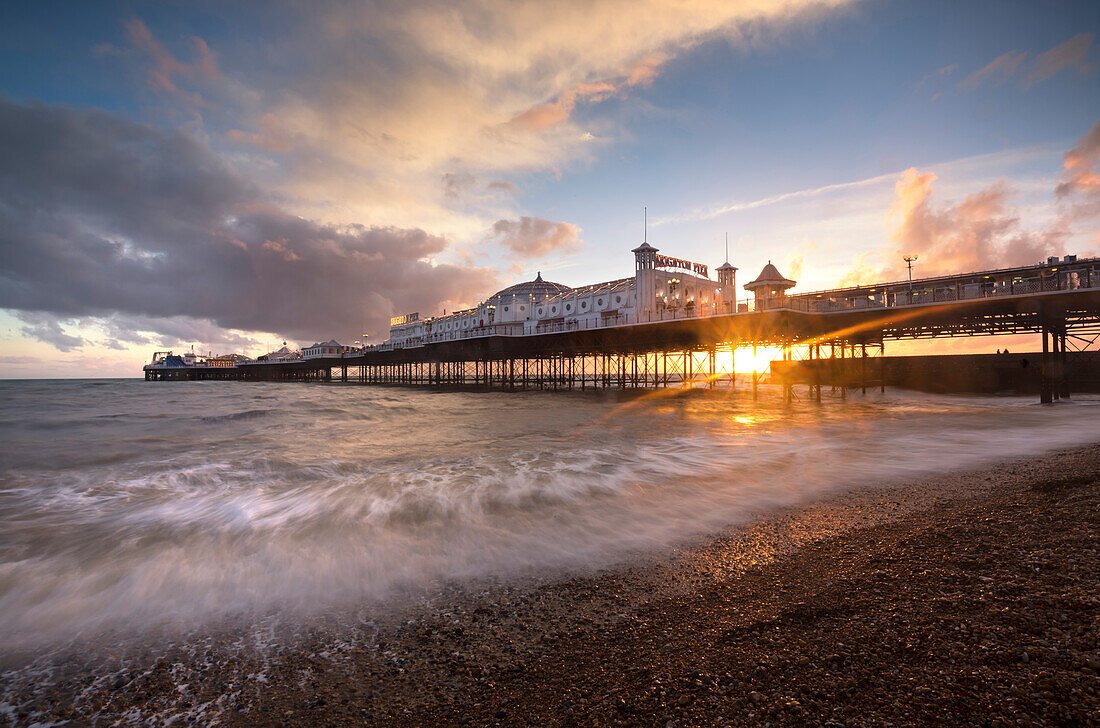Brighton Pier bei Sonnenuntergang mit dramatischen Himmel und Wellen den Strand spülen, Brighton, East Sussex, England, Großbritannien, Europa