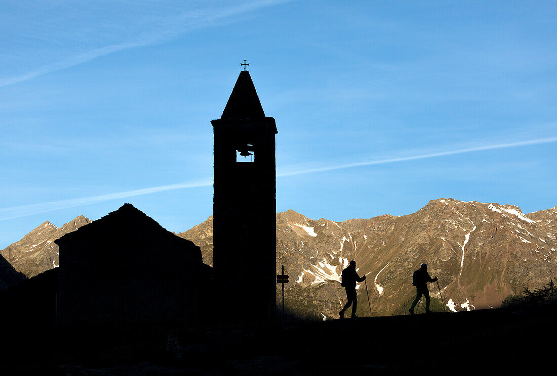 Silhouettes of hikers at the old church at dawn, San Romerio Alp, Brusio, Canton of Graubünden, Poschiavo valley, Switzerland