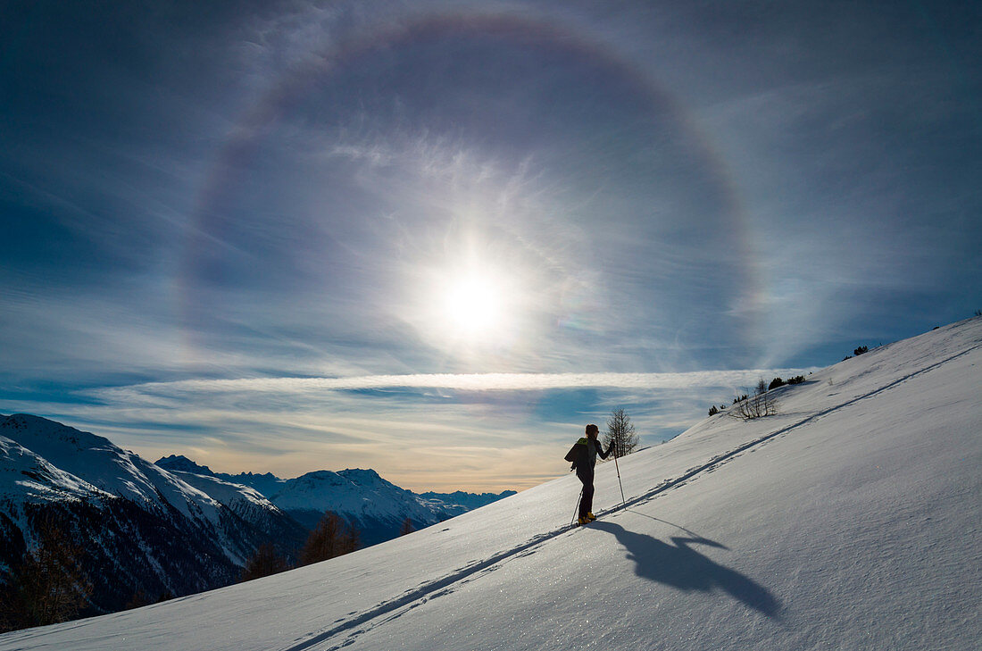 Skifahrer bergauf unter einem 22 ° Sonnenhalo im Winter, Muntuischè, Engadin, Schweiz