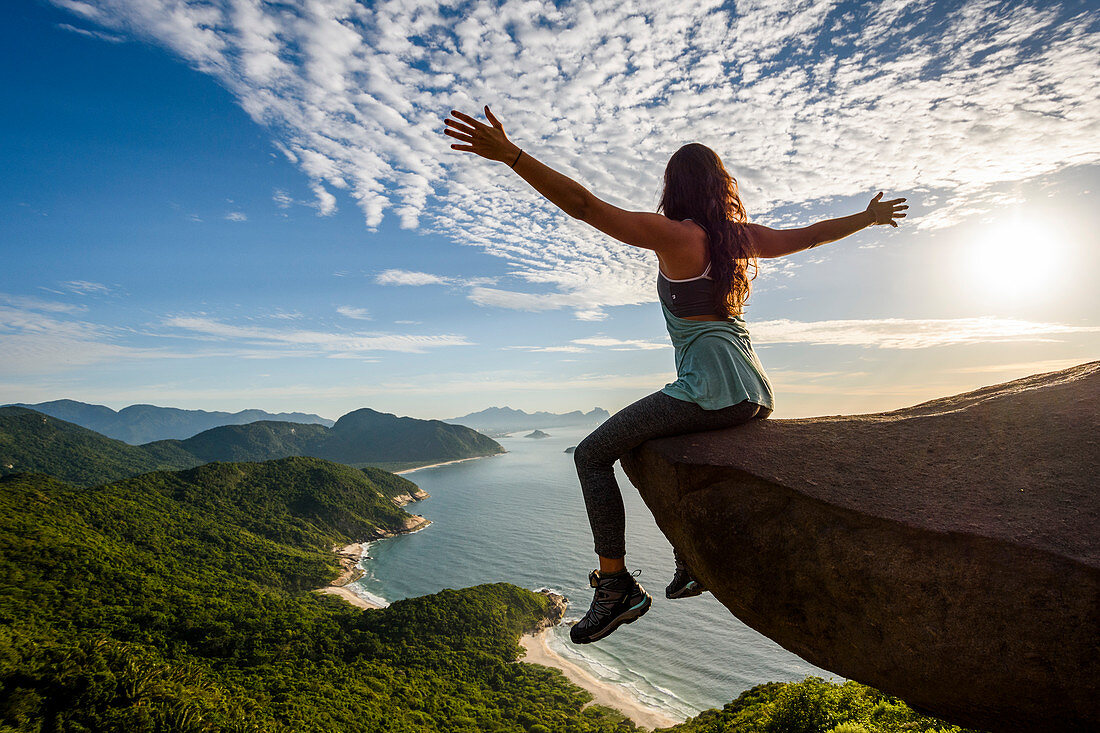 Woman sitting on the edge of the mountain with arms open in Pedra do TelÃ©grafo, Barra de Guaratiba, west side of Rio de Janeiro, Brazil