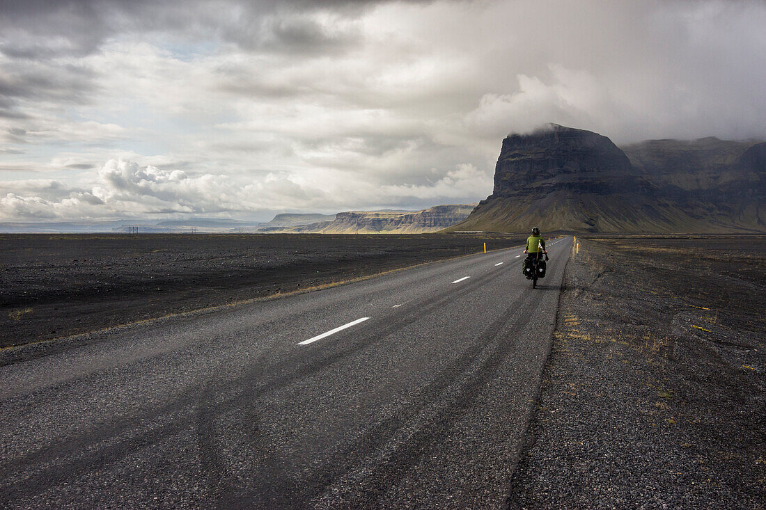 Woman riding touring bicycle on a straight road towards a mountain in a volcanic landscape.