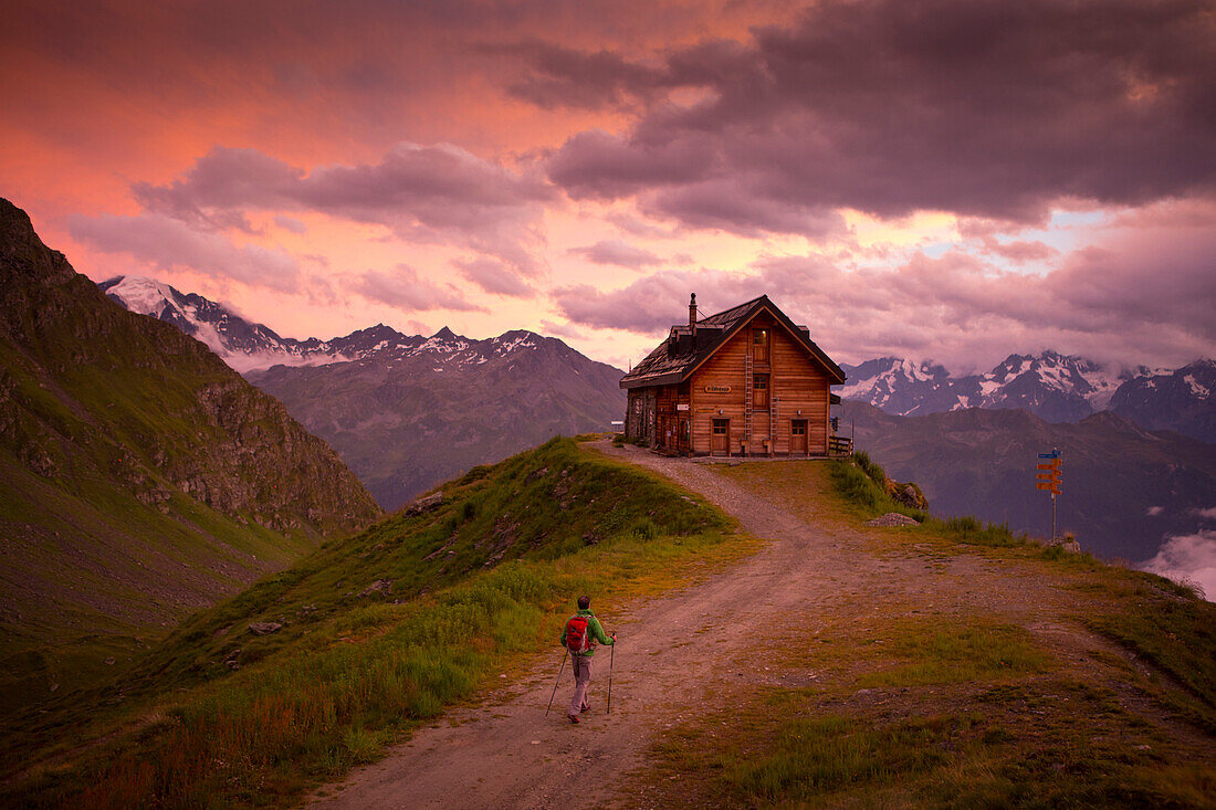 A hiker is almost at the Cabane du Mont Fort, a mountain hut near Verbier. The sunset puts the sky in pink and purple colors. It rained all day, and cleared just before this picture. The hut is one of the places hikers stay at during the Haute Route, a cl