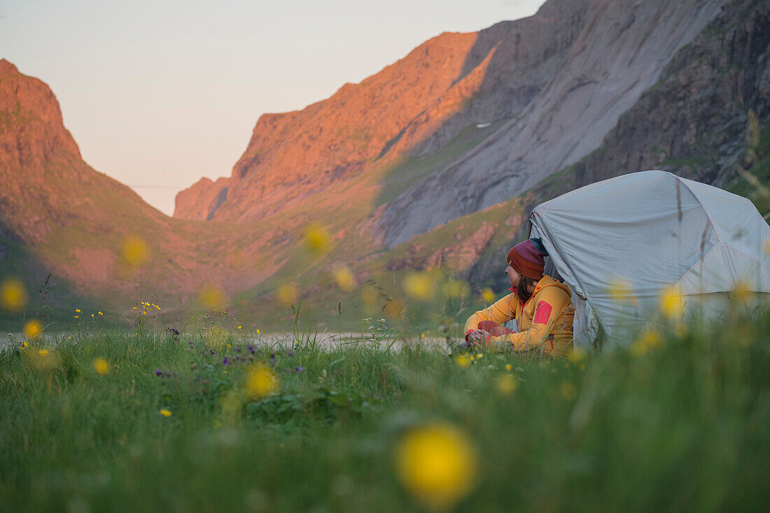 Weiblicher Wanderer sitzt Außenzelt umgeben von Sommerwildblumen, Horseid-Strand, Moskenesøy, Lofoten-Inseln, Norwegen