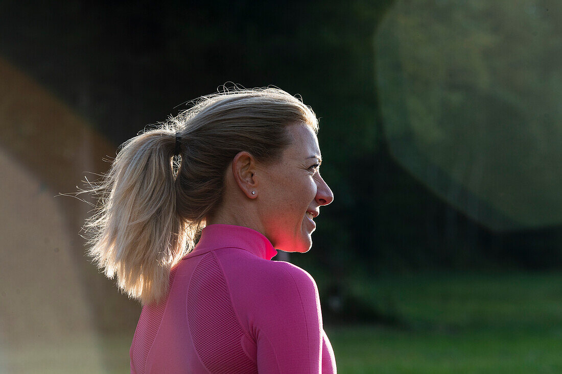 close up of a blond woman in profile at sunset, wearing a pink top and forest in the blurred background