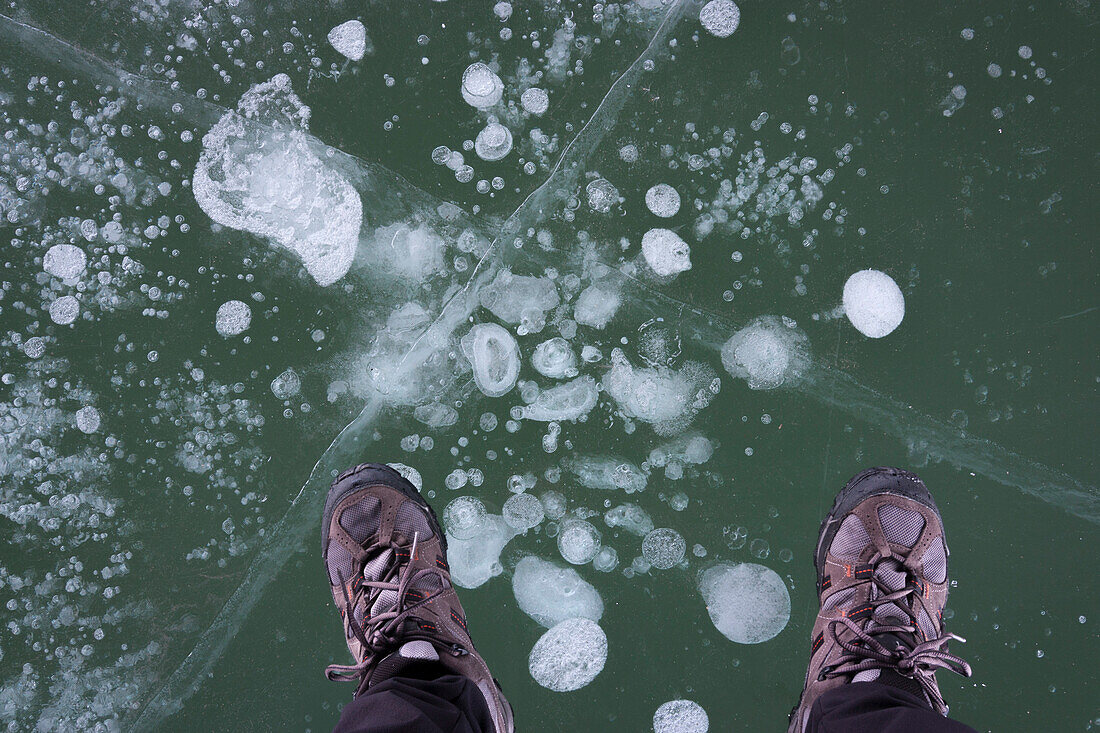 Der Mann steht auf durchscheinendem, rissigem Eis, der Wanderschuhe trägt, nur Füße sind sichtbar.