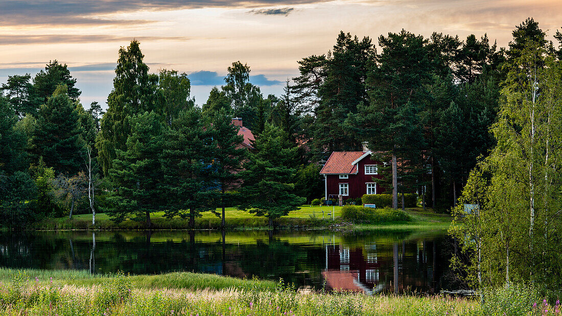 typisches schwedisches Haus, das in den Gewässern eines Sees nahe Mora in Schweden im Sommer mit Bäumen ringsum und schönem Himmel mit Wolken und netten Sonnenunterganglicht reflektiert.